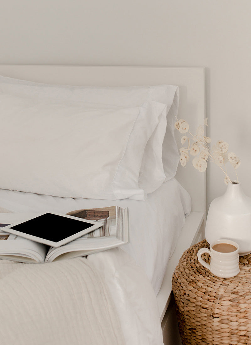Un ipad apagado apoyado sobre una cama. Al lado de la cama se encuentra una taza de café sobre una mesa.