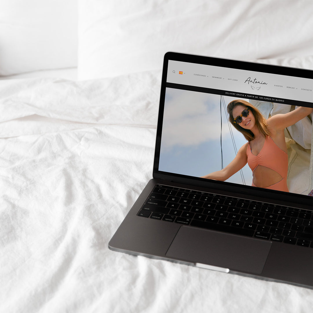 Una laptop abierto sobre una cama. En su pantalla se encuentra abierto el inicio de la página web de la marca Antonia.