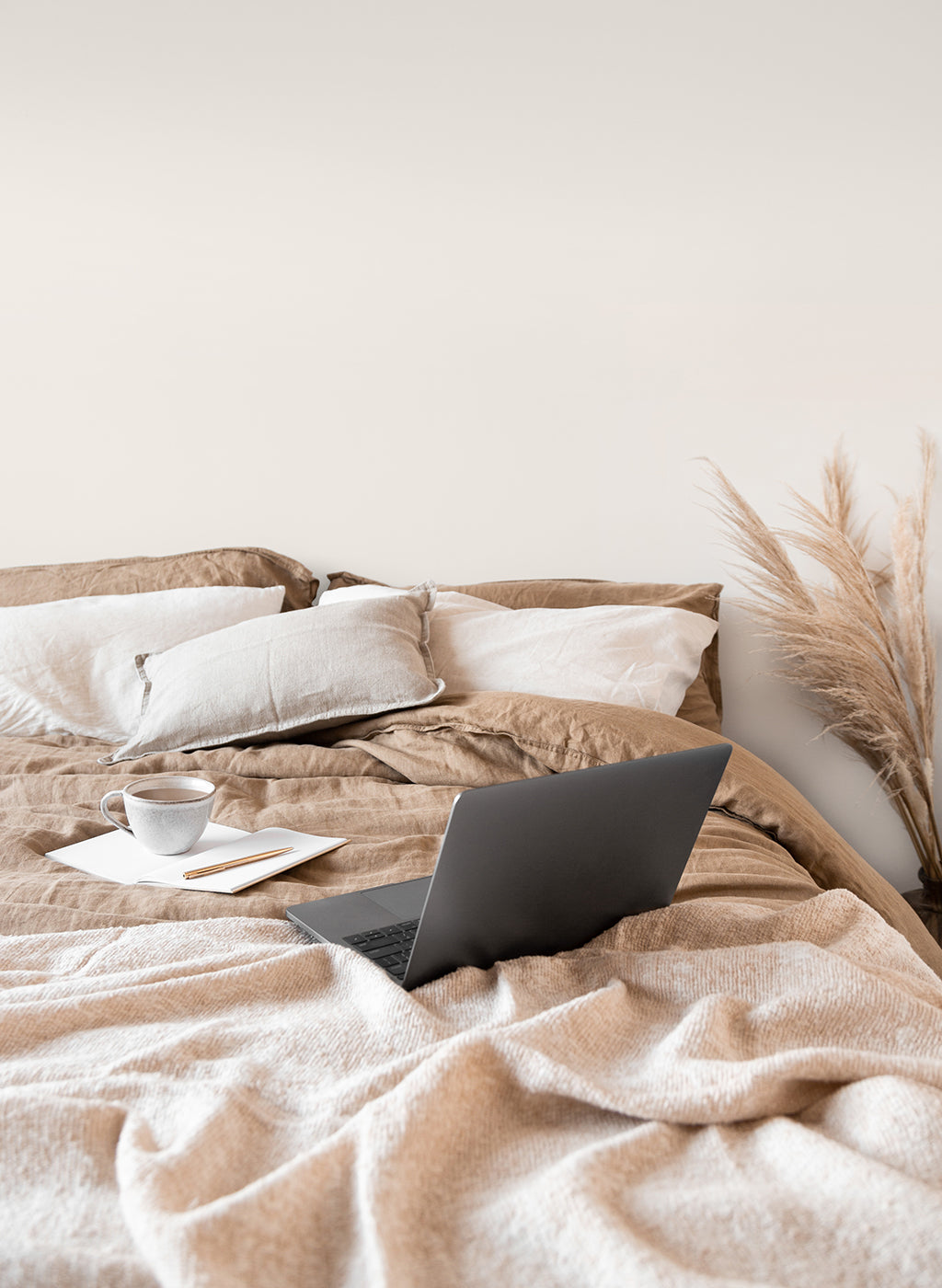 Laptop abierta sobre una cama. A su lado se encuentra una taza de café apoyada sobre un cuaderno. 