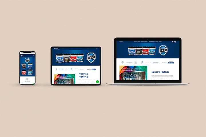 Un Iphone, un Ipad y una laptop mostrando cada uno el inicio de la página web de Pinturas Índigo. Los tres dispositivos están uno al lado del otro sobre un fondo color entero beige. 